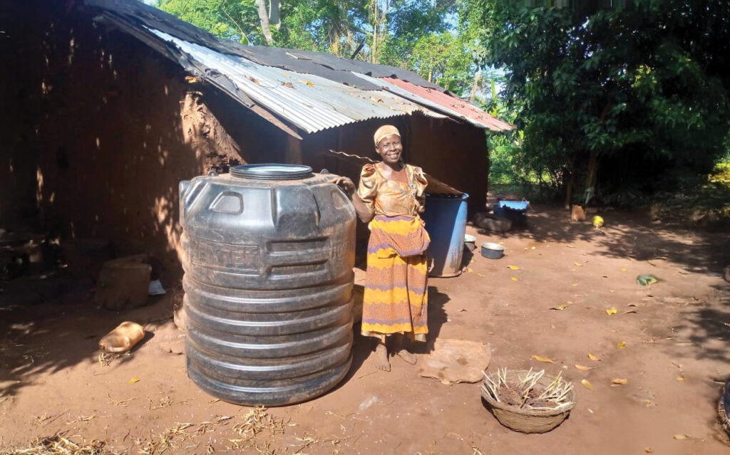 Justine is standing with a beaming smile beside her rainwater harvesting tank outside her home.