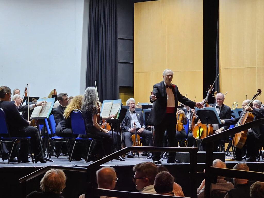 Darrell Davison faces the audience and speaks, with the orchestra waiting to start behind him