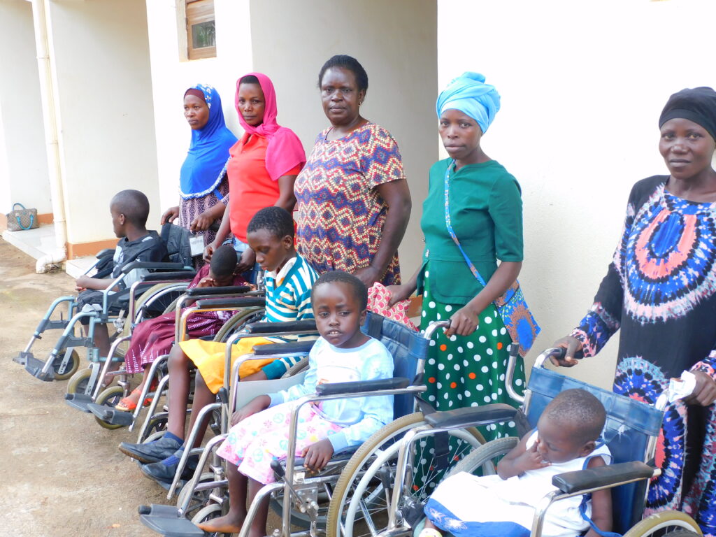 Five mothers stand in line holding their children's wheelchairs. They are wearing bright colours and patterns, and look directly at the camera