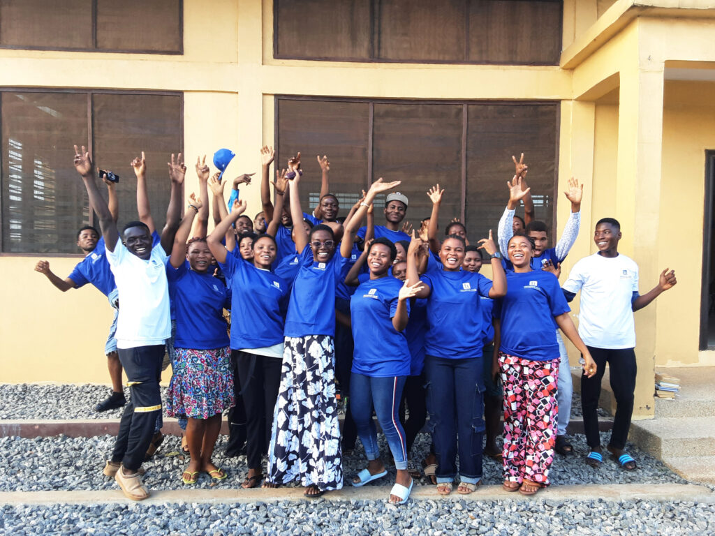 Around twenty students hold their hands in the air smiling and wearing IN Ghana t shirts