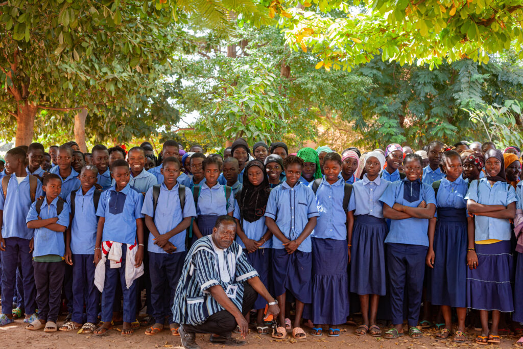 about 25 students in blue and navy uniform gather together under the trees with the ED crouching down and smiling in front of them