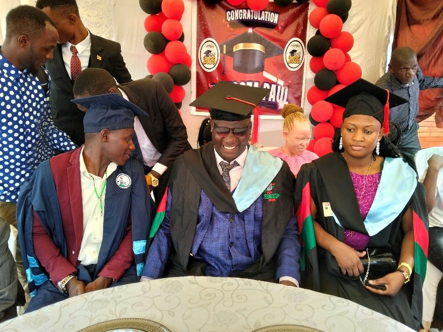 With balloons in the backdrop and wearing graduation robes and caps, Kirabo laughs between two friends at graduation day