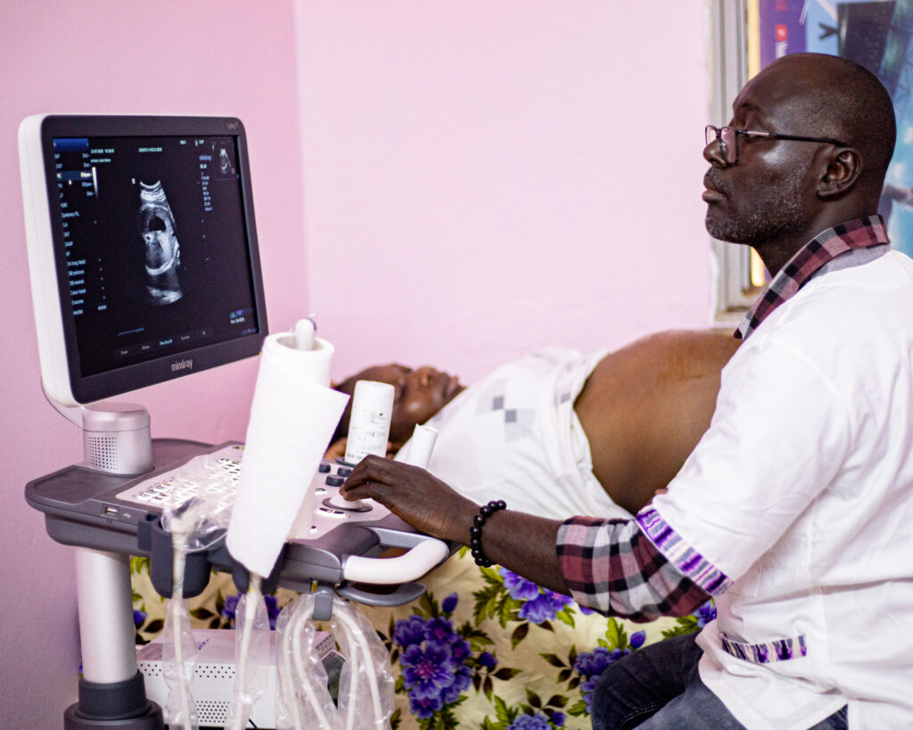 A doctor looks at the ultrasound scanner while a pregnant lady lies down behind him.