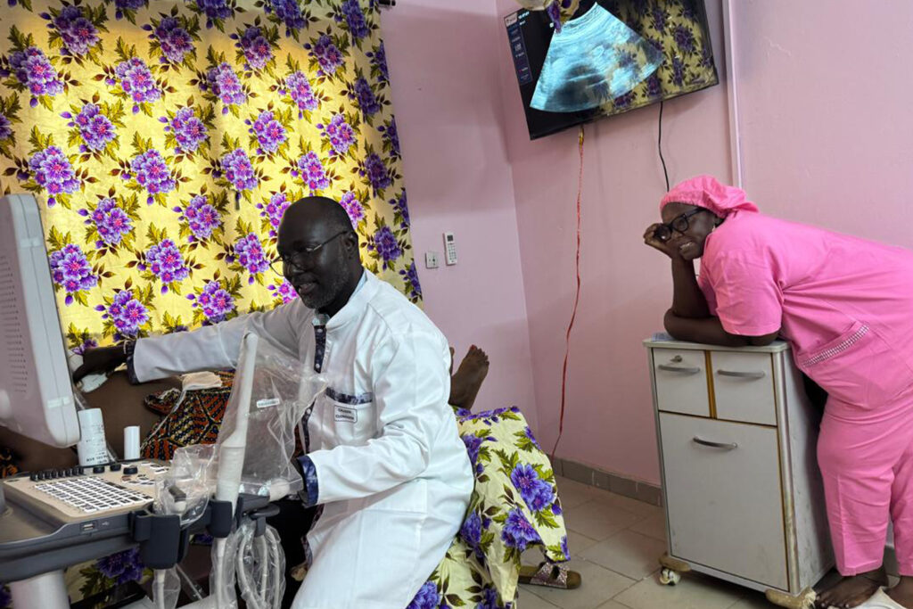 The doctor smiles as he uses the scanner and a nurse in pink scrubs smiles behind him